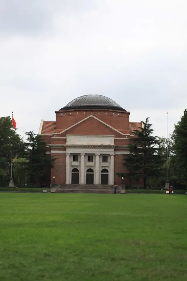 The domed red-brick Grand Auditorium of Tsinghua University in Beijing, seen across a wide green lawn under an overcast sky.