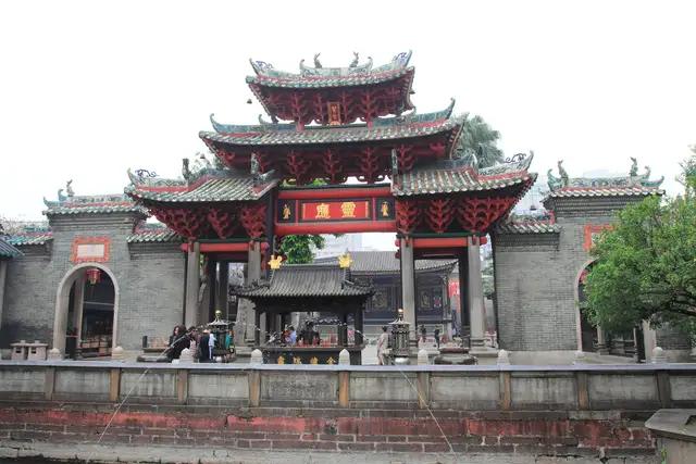 The Lingying memorial archway of the Foshan Ancestral Temple, with layered red brackets and green-glazed roofs, stands above a stone-walled pond under an overcast sky.