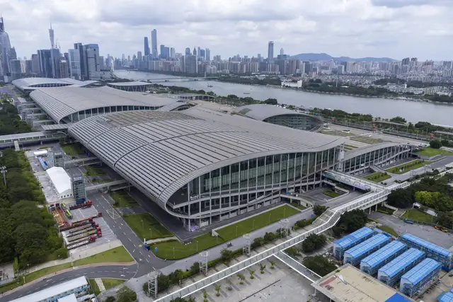 Aerial view of the curved silver roofs of the Canton Fair Complex on Pazhou Island beside the Pearl River, with the Guangzhou skyline in the distance.