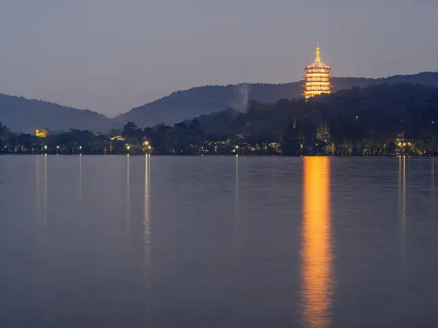 The illuminated Leifeng Pagoda on a wooded hill above West Lake in Hangzhou at dusk, its golden light reflected across calm water.