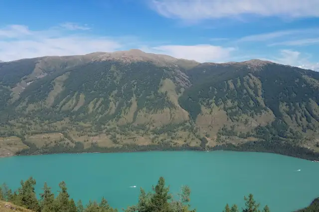 Jade-green Kanas Lake beneath forested mountain slopes under a blue summer sky, Altay, Xinjiang