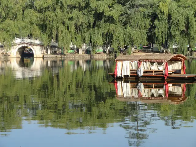 A covered wooden boat moored on the calm water of Green Lake Park in Kunming, with willow trees and a white stone arch bridge behind.