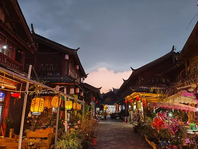 A stone-paved street in Lijiang old town at dusk, lined with wooden houses, yellow lanterns and flower stalls under a grey evening sky.