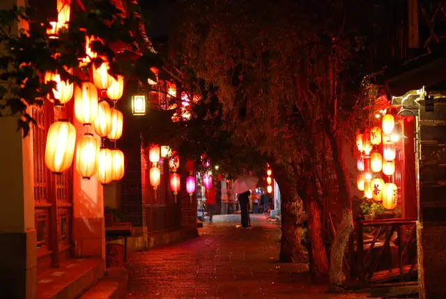 A lane in Lijiang old town on a rainy night, glowing red and orange from clusters of paper lanterns hung along the wooden houses.