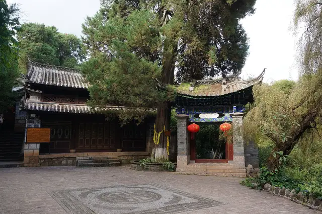 A quiet stone-paved courtyard at Longquan Temple in Shuhe old town, Lijiang, with an old cypress tree, a tiled gateway and red lanterns.