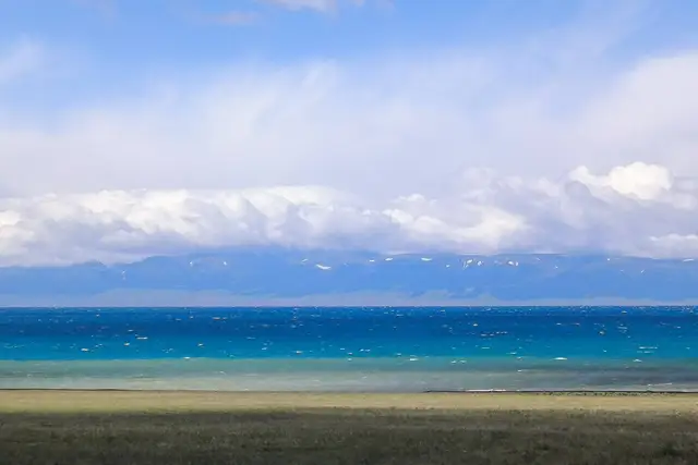 Turquoise water of Sayram Lake under a band of white cloud, with snow-flecked Tianshan mountains beyond and a grassy shore in front