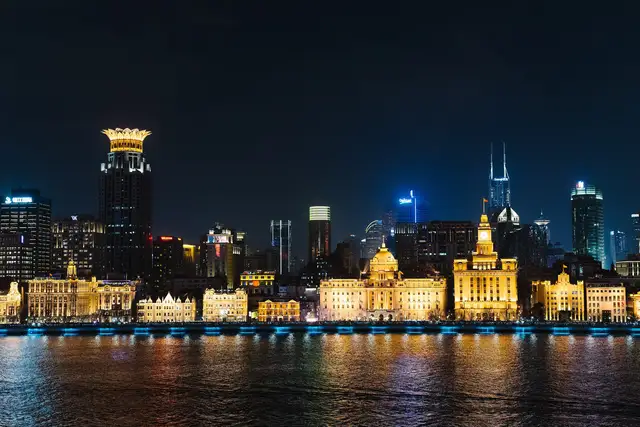 The illuminated historic facades of the Bund in Shanghai line the Huangpu River at night, with modern towers rising behind them.