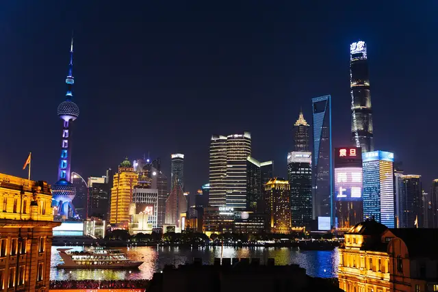 The Lujiazui skyline in Shanghai at night, with the Oriental Pearl Tower and Shanghai Tower lit up across the Huangpu River and Bund rooftops in the foreground.