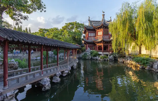 A red two-storey pavilion with upturned eaves, a roofed walkway over the pond and a weeping willow in Yu Garden, Shanghai, in late-afternoon light.