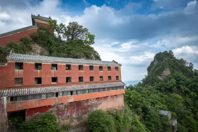 A red-walled Taoist palace perched on a cliff in the Wudang Mountains, with green peaks and clouds beyond