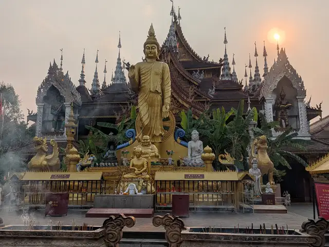 A golden standing Buddha statue in front of the dark spired roofs of Zongfo Temple in Jinghong, Xishuangbanna, with the sun low in a hazy sky.